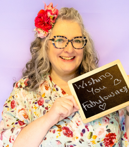 Vie is smiling with flowers in her hair, wearing a floral dress and holding up a chalkboard which says"Wishing you fabulousness"