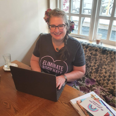 Vie is sat at a table,smiling at the camera and typing on her laptop