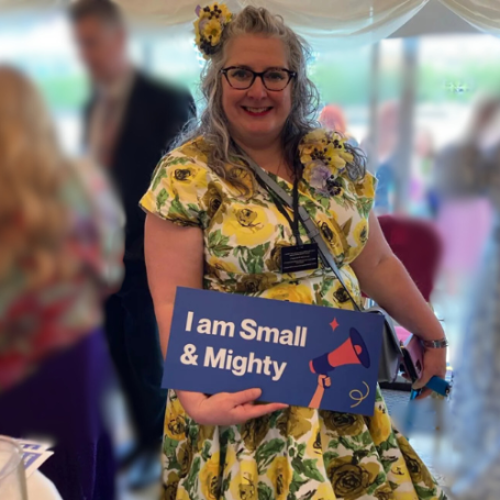 Vie holding a large card with "I am small and mighty" written on it. Vie is wearing a yellow flowery dress and yellow flower hair decoration