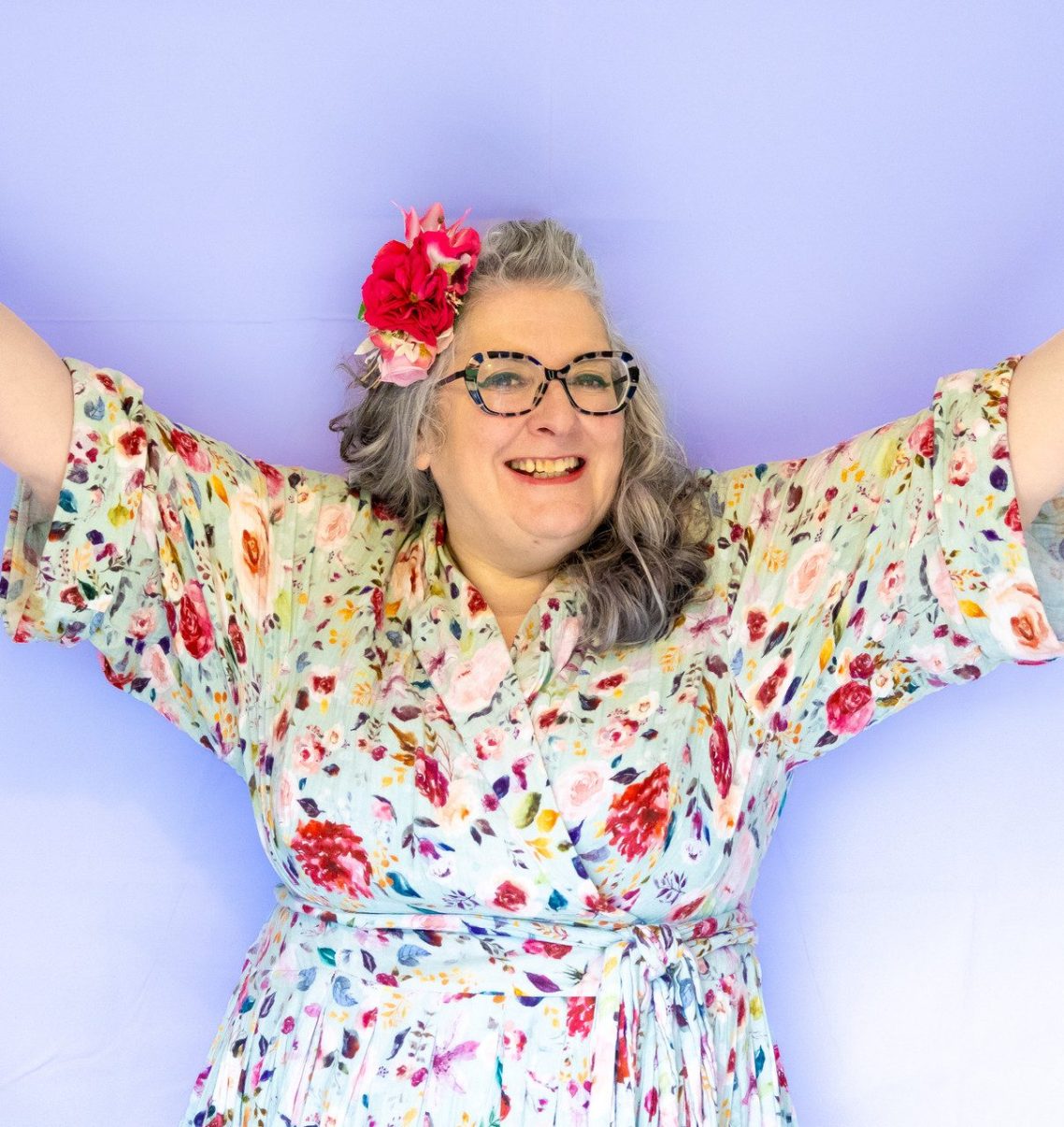 Smiling woman with grey hair and glasses, wearing a floral robe, with arms raised.